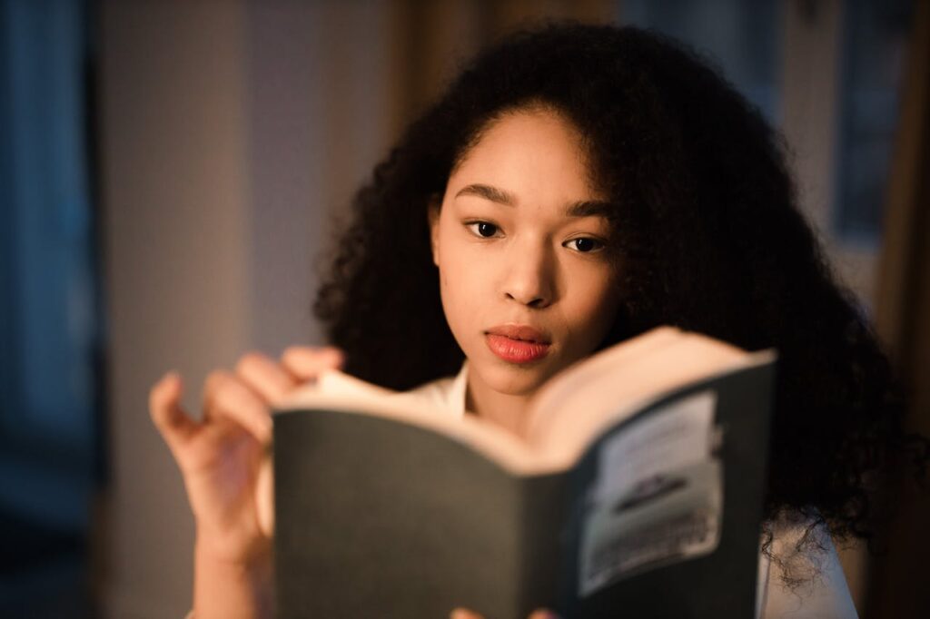 A young woman with curly hair is engrossed in reading a book indoors.